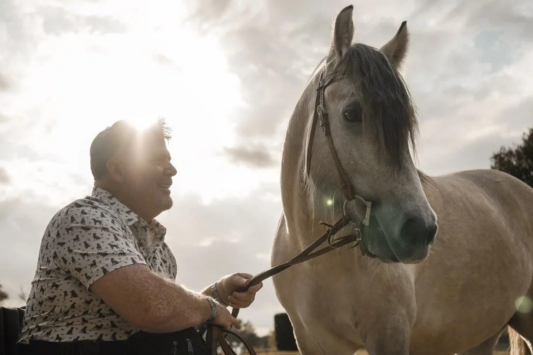 A man with dark hair and a patterned shirt smiling at a white horse with black mane outdoors during daytime with cloudy sky.
