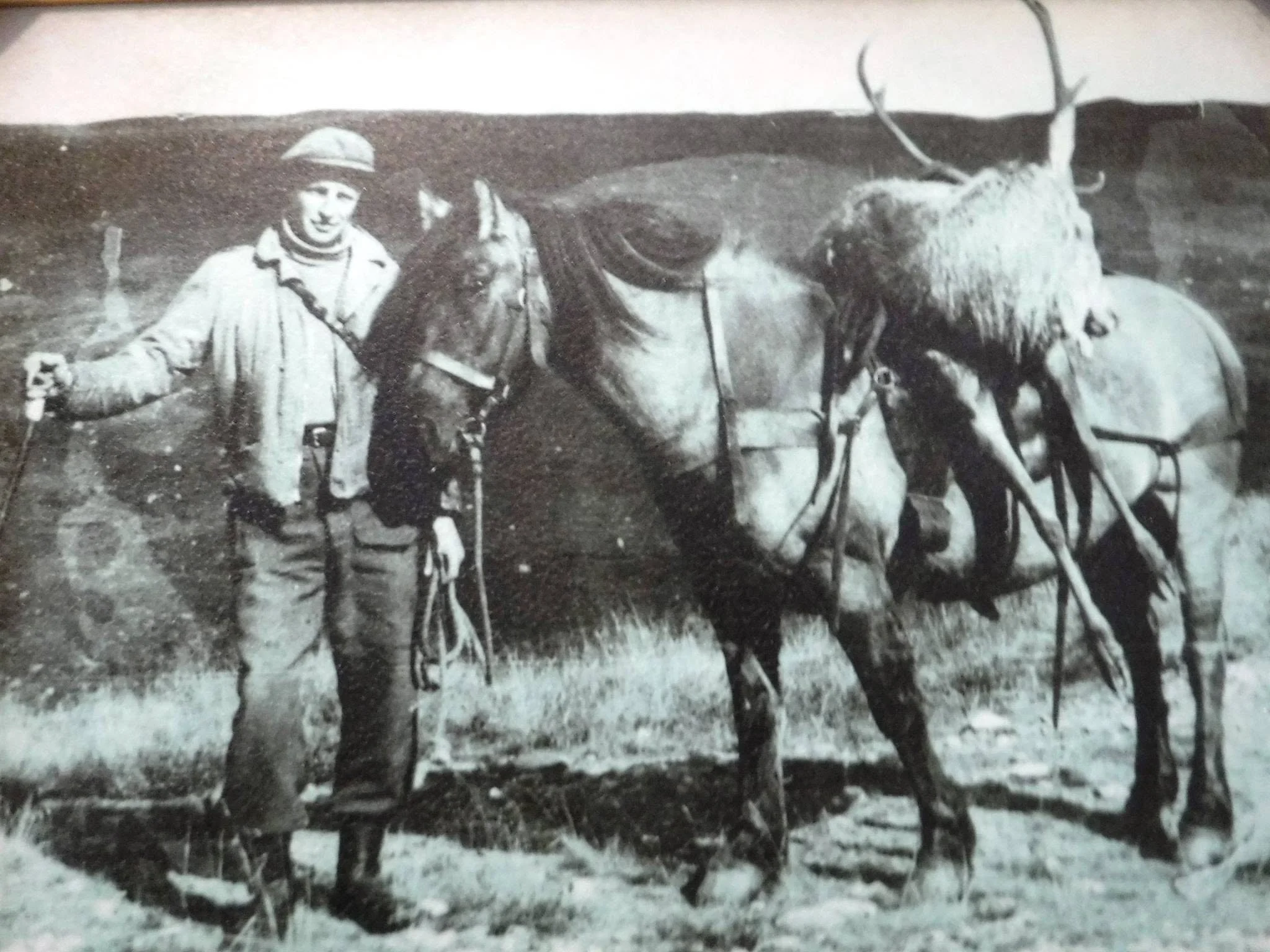 Black-and-white photo of a man standing next to two reindeer in an outdoor landscape.