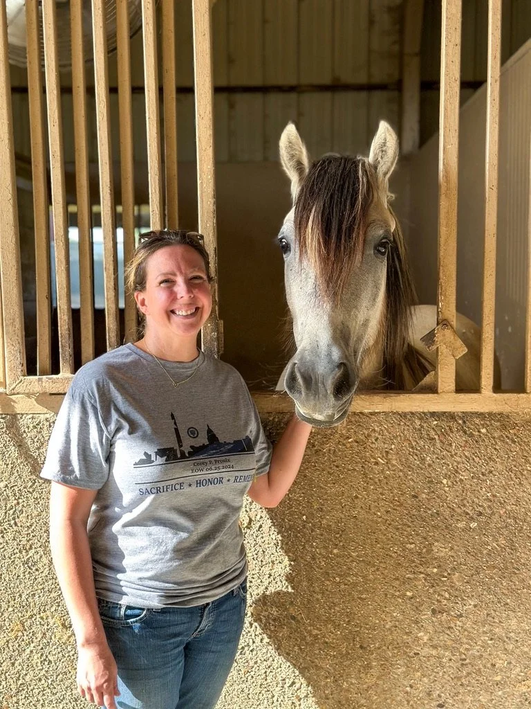 A woman smiling and touching a gray horse in a stable.