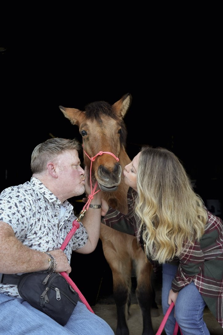 A couple kissing a horse's nose while holding its halter, with the horse's head between them and the dark background.