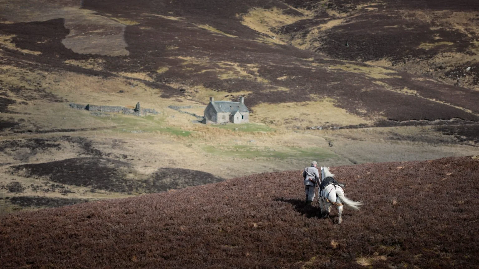 A person walking a white horse on a brown hillside with a stone house in the background, set in a rural landscape.