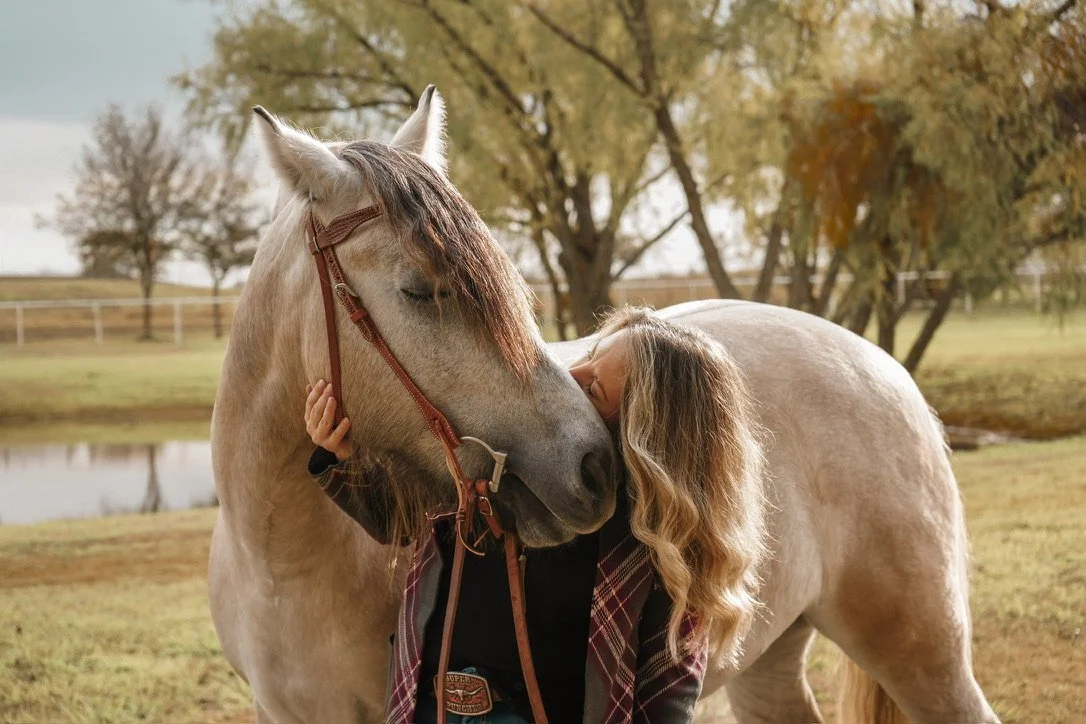 A girl hugging a white horse outdoors near a pond, with trees showing fall foliage in the background.