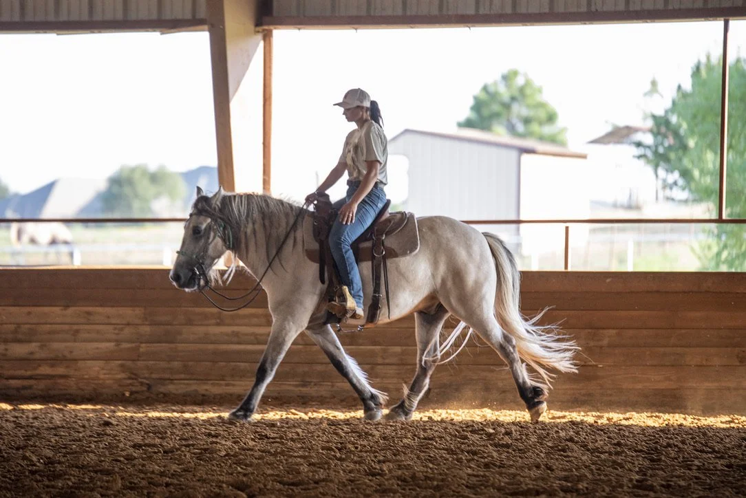 A woman riding a light-colored horse inside an indoor riding arena.
