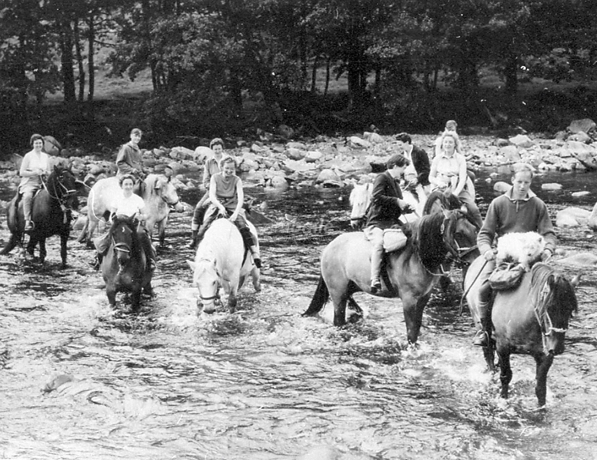 A black and white photo of a group of people riding horses across a shallow river, with trees and forest in the background.