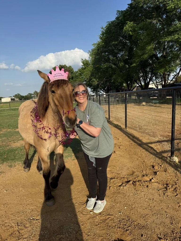 Woman standing next to a decorated miniature horse wearing a pink birthday crown, both outdoors on a sunny day near a fence and trees.
