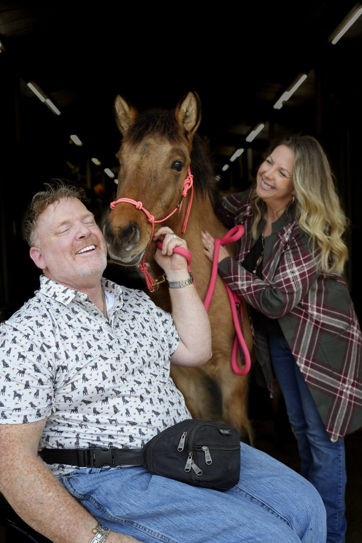 A smiling man sitting and a woman standing behind a brown horse with a pink halter, both touching the horse in a stable or barn with overhead lights.