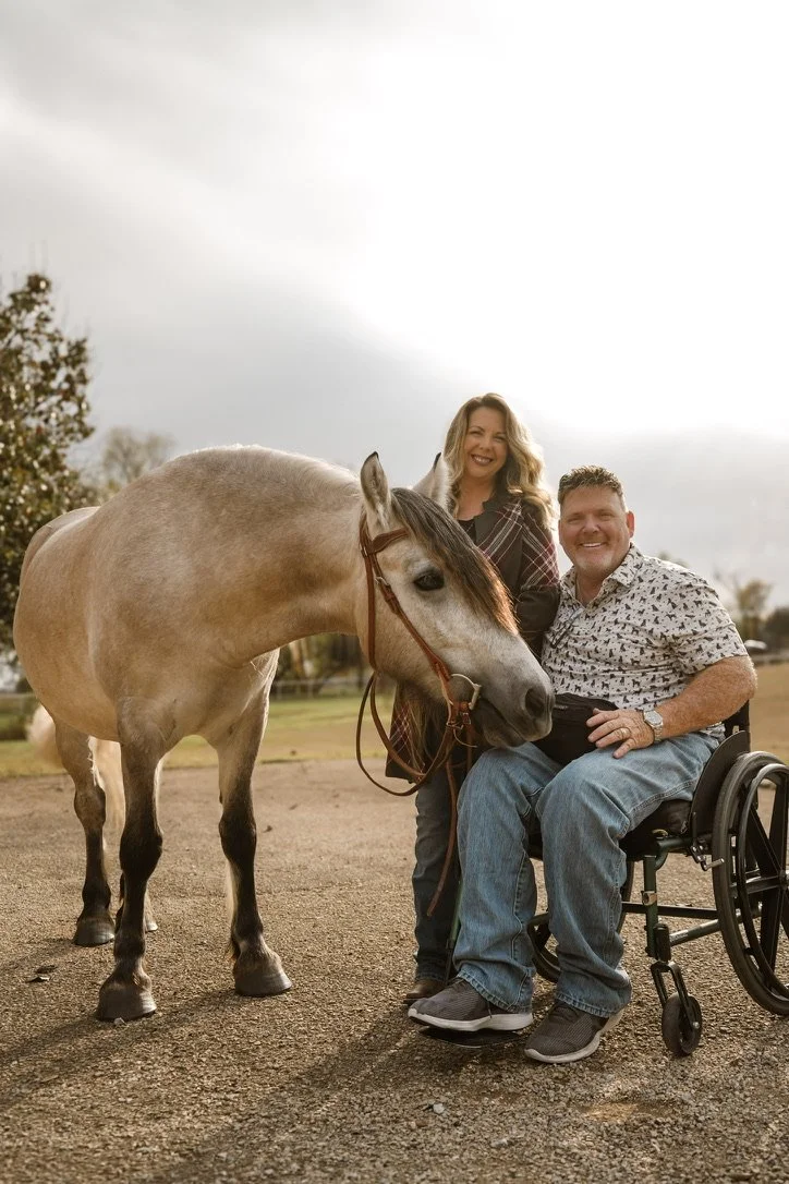 A woman standing next to a man in a wheelchair, both smiling and holding a horse's bridle, outdoors on a cloudy day.