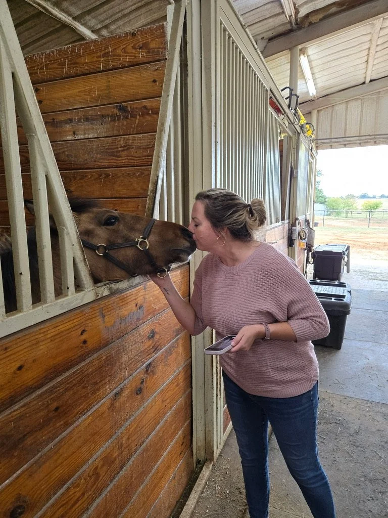 A woman with brown hair in a bun wearing a pink sweater and blue jeans is hugging and kissing a horse's nose through the stable window.
