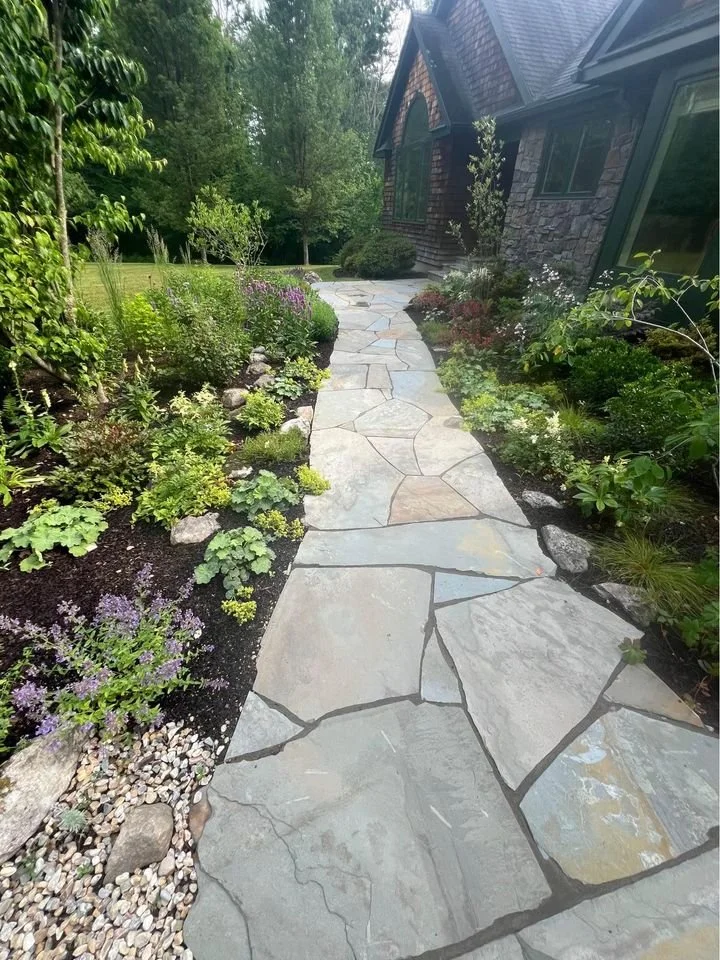 A stone walkway leading past a garden with various green plants and flowers, next to a house with a dark brown exterior and chimney, surrounded by trees and lush greenery.