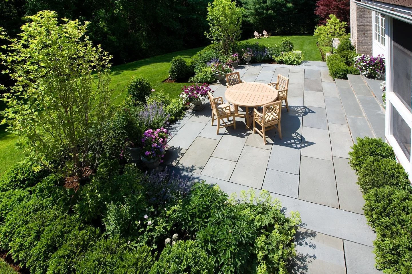 A backyard patio with a round wooden table and six matching chairs, surrounded by lush green bushes and flowering plants, leading to a grassy lawn with trees in the background.