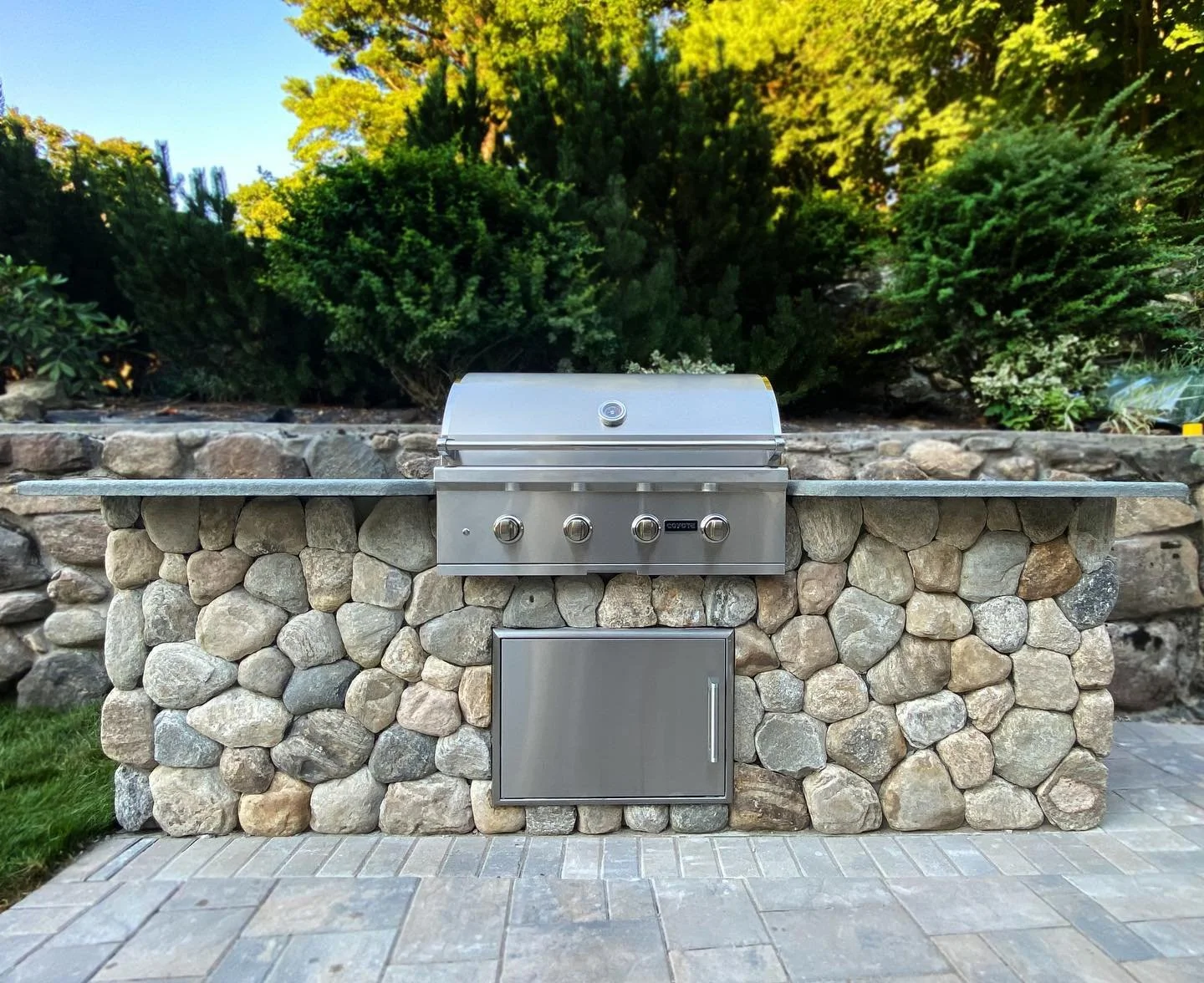 Outdoor stainless steel grill on stone wall with greenery in background.