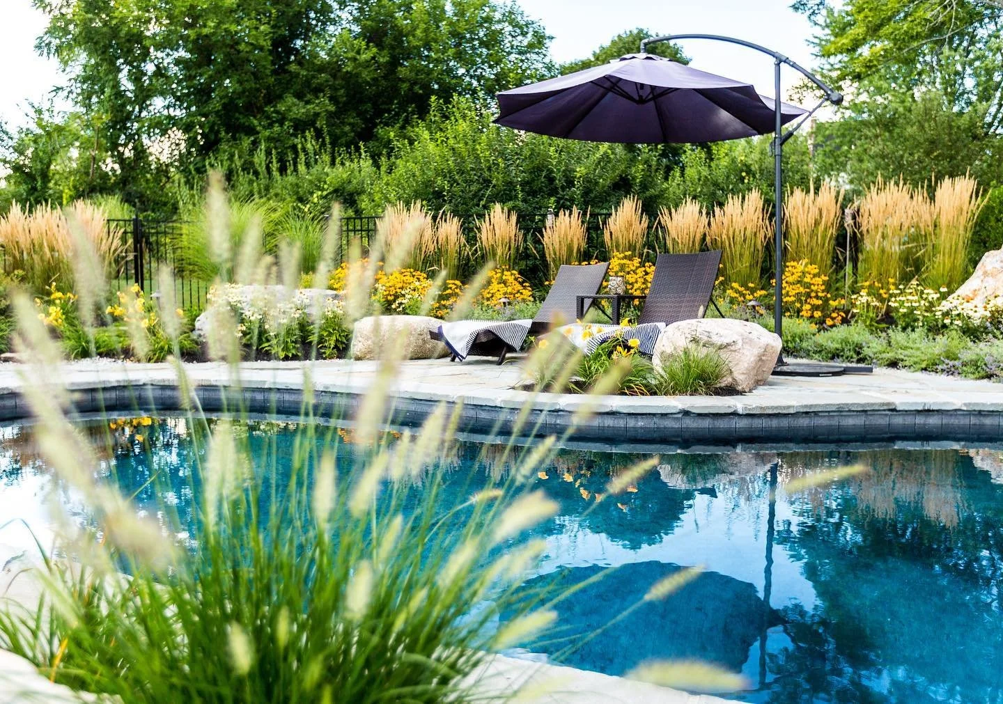 A backyard swimming pool area with poolside chairs, a large umbrella, and colorful flower landscaping.