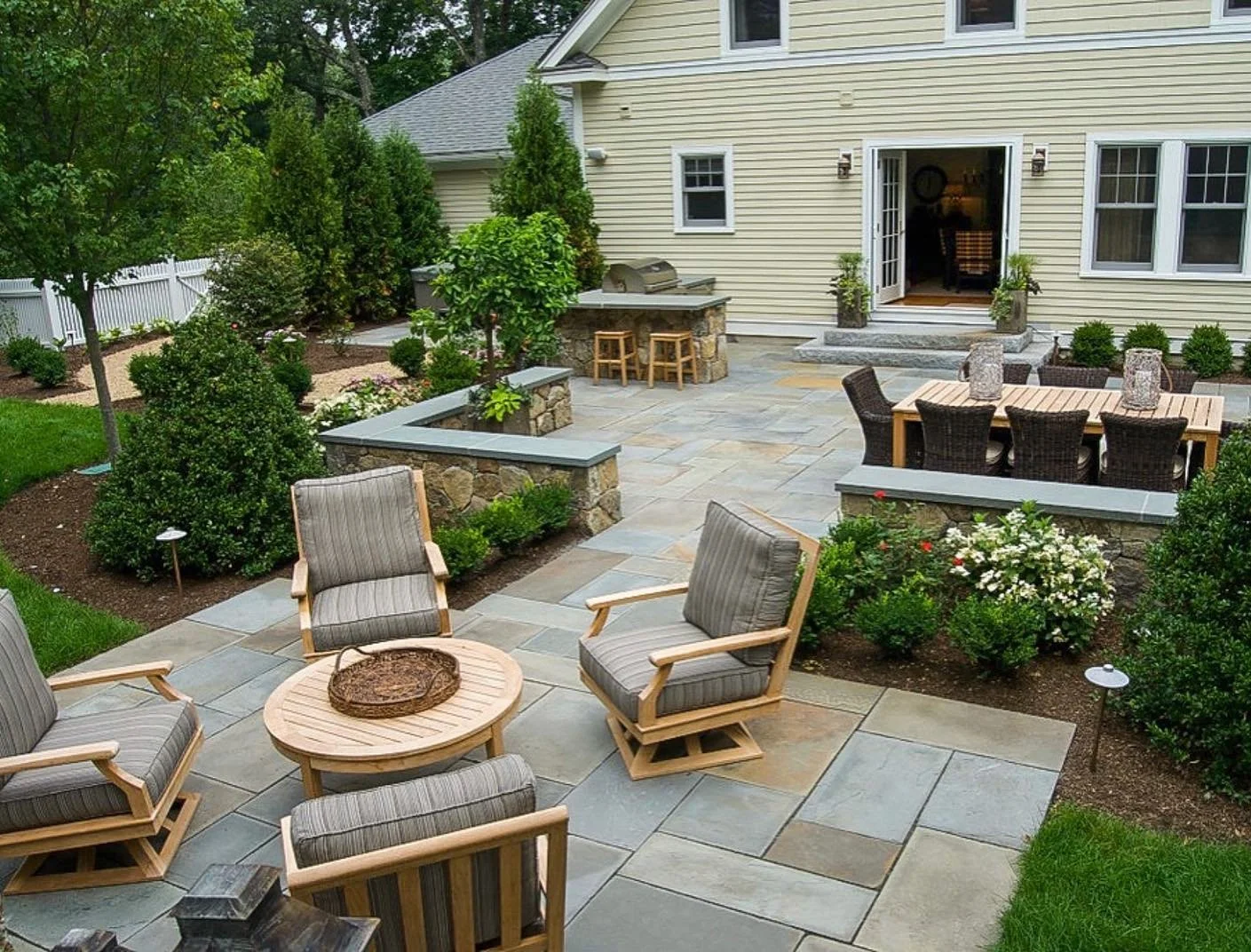 Backyard patio with outdoor seating, a dining table, a built-in grill, and lush green plants surrounding a beige house.