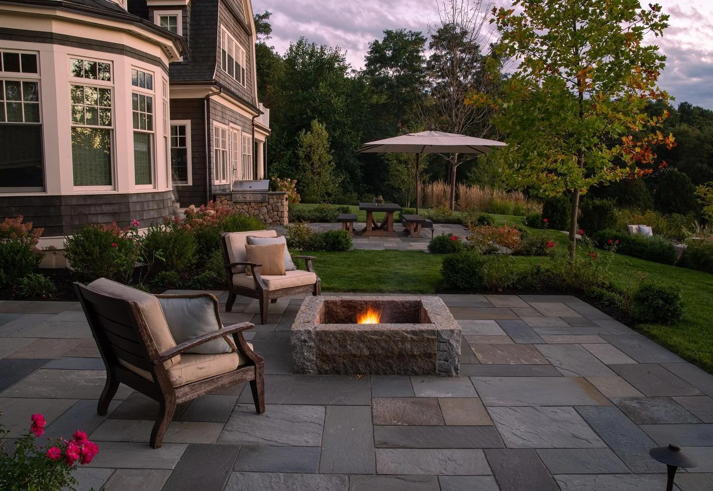 A backyard patio at sunset with four chairs surrounding a square stone fire pit, an umbrella-shaded dining table, green grass, flowering bushes, and a large tree.