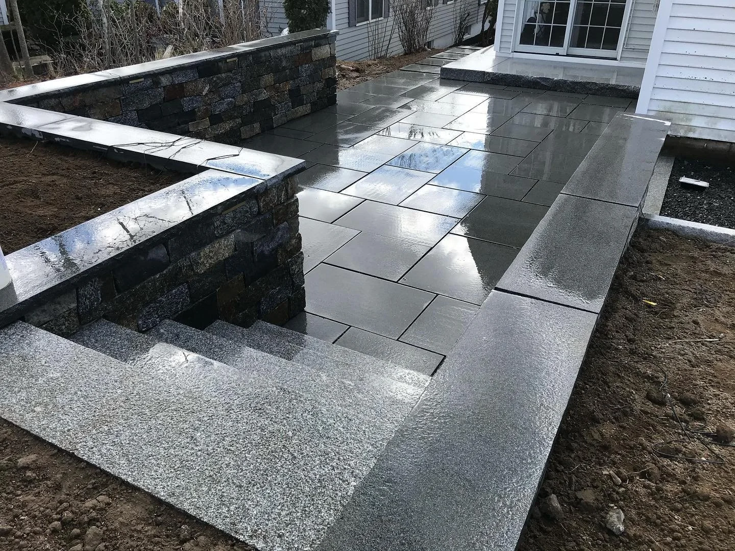 Recently installed grey stone patio with matching steps and a stone wall, wet from rain, located outside a house with white siding and a sliding glass door.