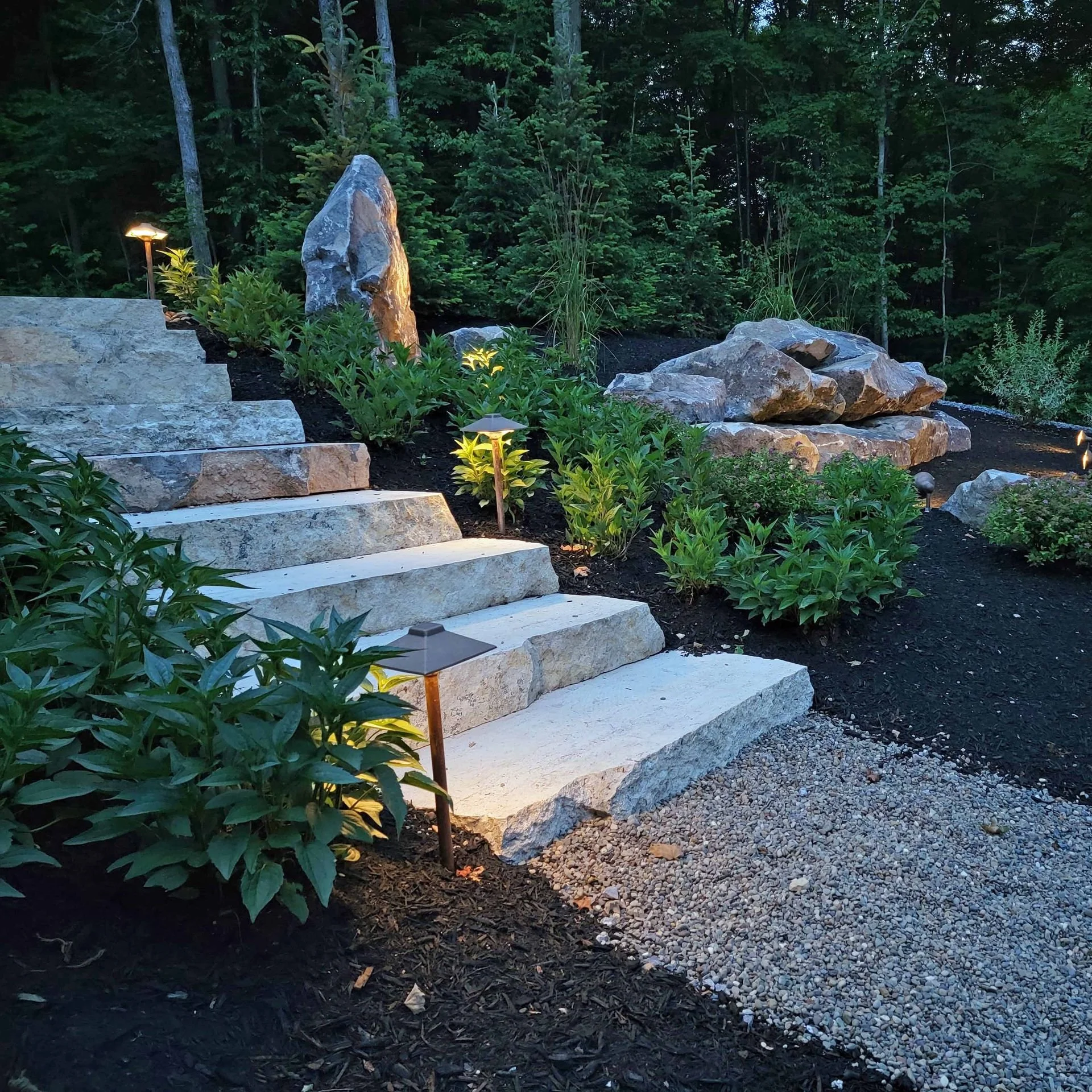 Nighttime garden scene with stone steps, large rocks, greenery, small trees, and landscape lighting.