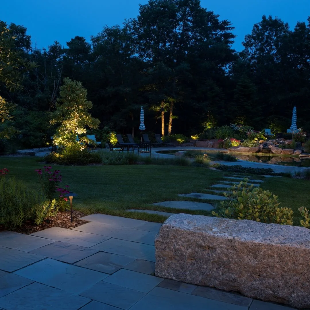 A backyard at dusk with a stone patio, landscaped garden, lit trees, lounge chairs, and a swimming pool surrounded by rocks and lush greenery.
