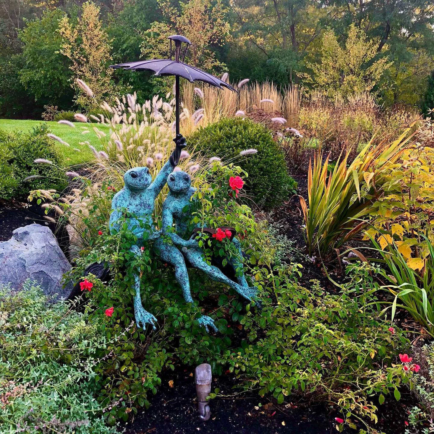 Garden scene with a metal sculpture of two frogs sitting on a bench, one frog holding an umbrella over the other, surrounded by various flowers and lush foliage.