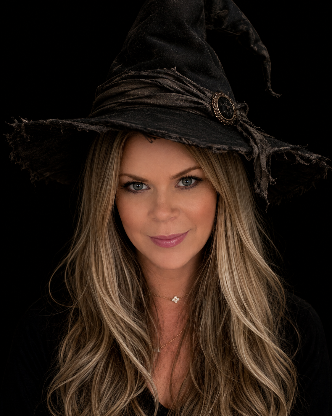 A woman with long blonde hair wearing a distressed black witch's hat with a silver buckle, and a black top, against a black background.