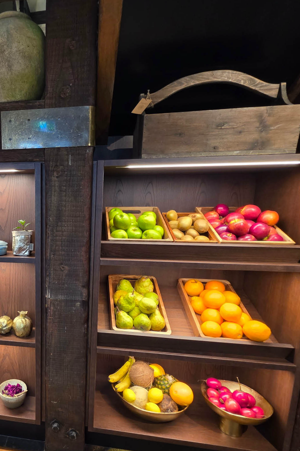 Wooden shelves with baskets of fresh fruits including green apples, yellow pears, oranges, bananas, and red apples, in a rustic setting.