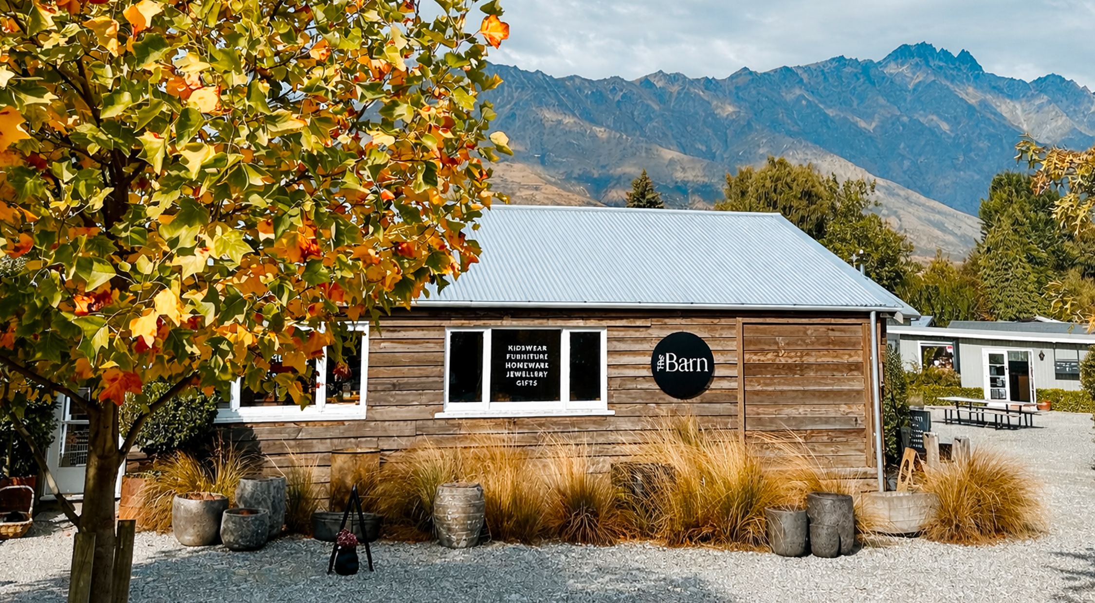 A small wooden shop with a sign that reads 'The Barn' and offers kid's wear, furniture, homeware, jewelry, and gifts. There is a colorful tree with orange, yellow, and green leaves in the foreground. Plastic planters and gravel ground surround the shop, with a mountain range and partly cloudy sky in the background.