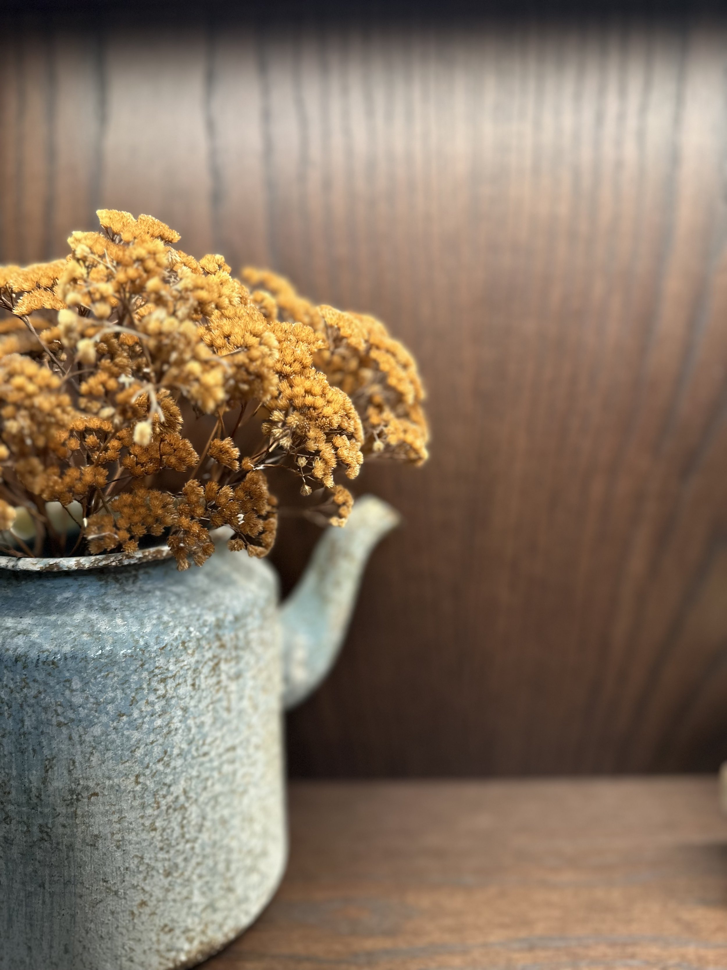A rustic ceramic teapot filled with dried yellow flowers on a wooden surface with a wooden wall background.