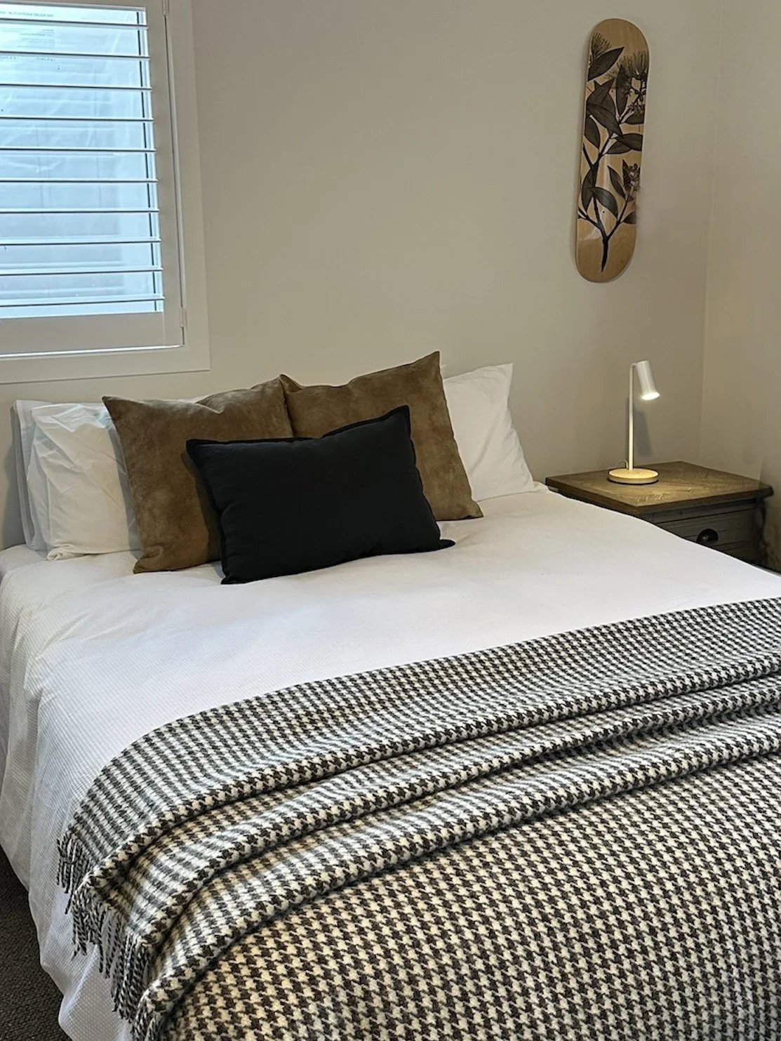 A neatly made bed with white bedding, four decorative pillows in different shades of brown and black, a black and white houndstooth throw blanket at the foot of the bed, all sourced in Queenstown from The Barn at Country Lane
