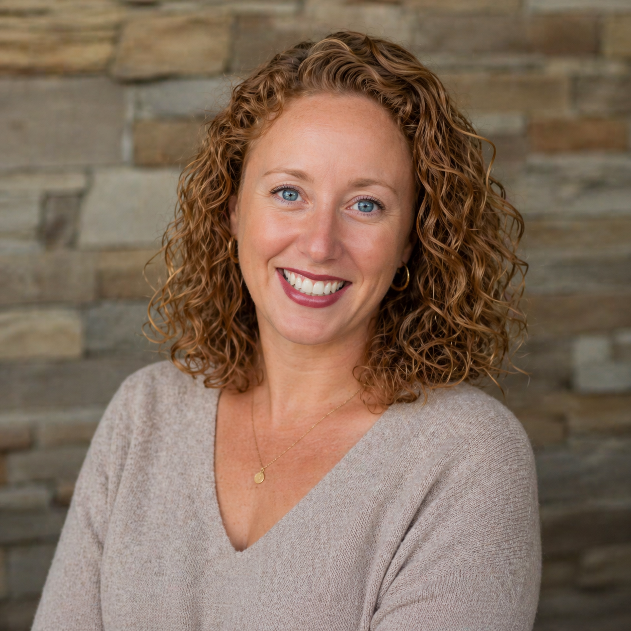 A woman with curly red hair smiling, wearing a beige top and a delicate gold necklace, against a stone wall background.