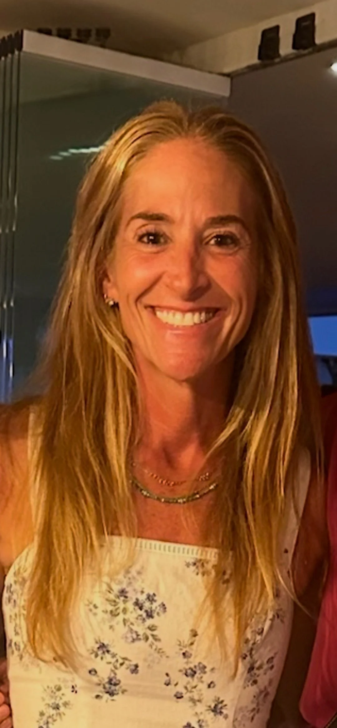 A smiling woman with long blonde hair, wearing a cream-colored floral dress and layered necklaces, posing indoors.