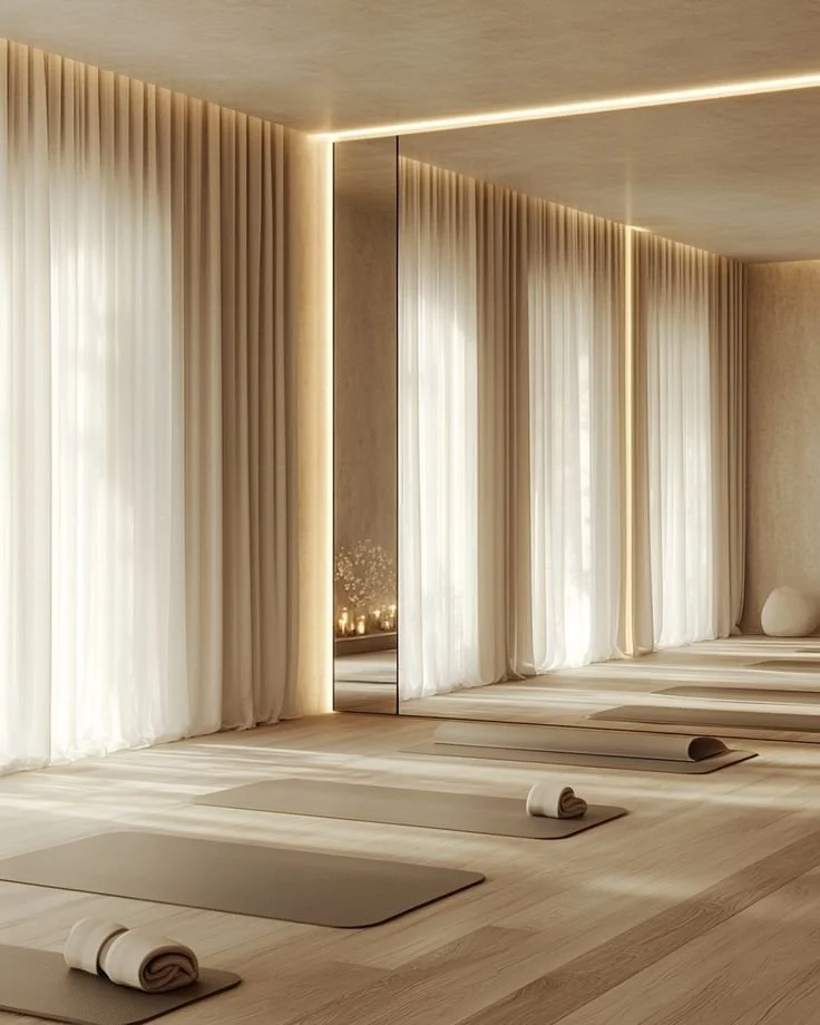 A minimalistic yoga studio with light wood floors, beige curtains, and large windows letting in natural light. Yoga mats with rolled towels are aligned on the floor, and a mirror is on one wall.