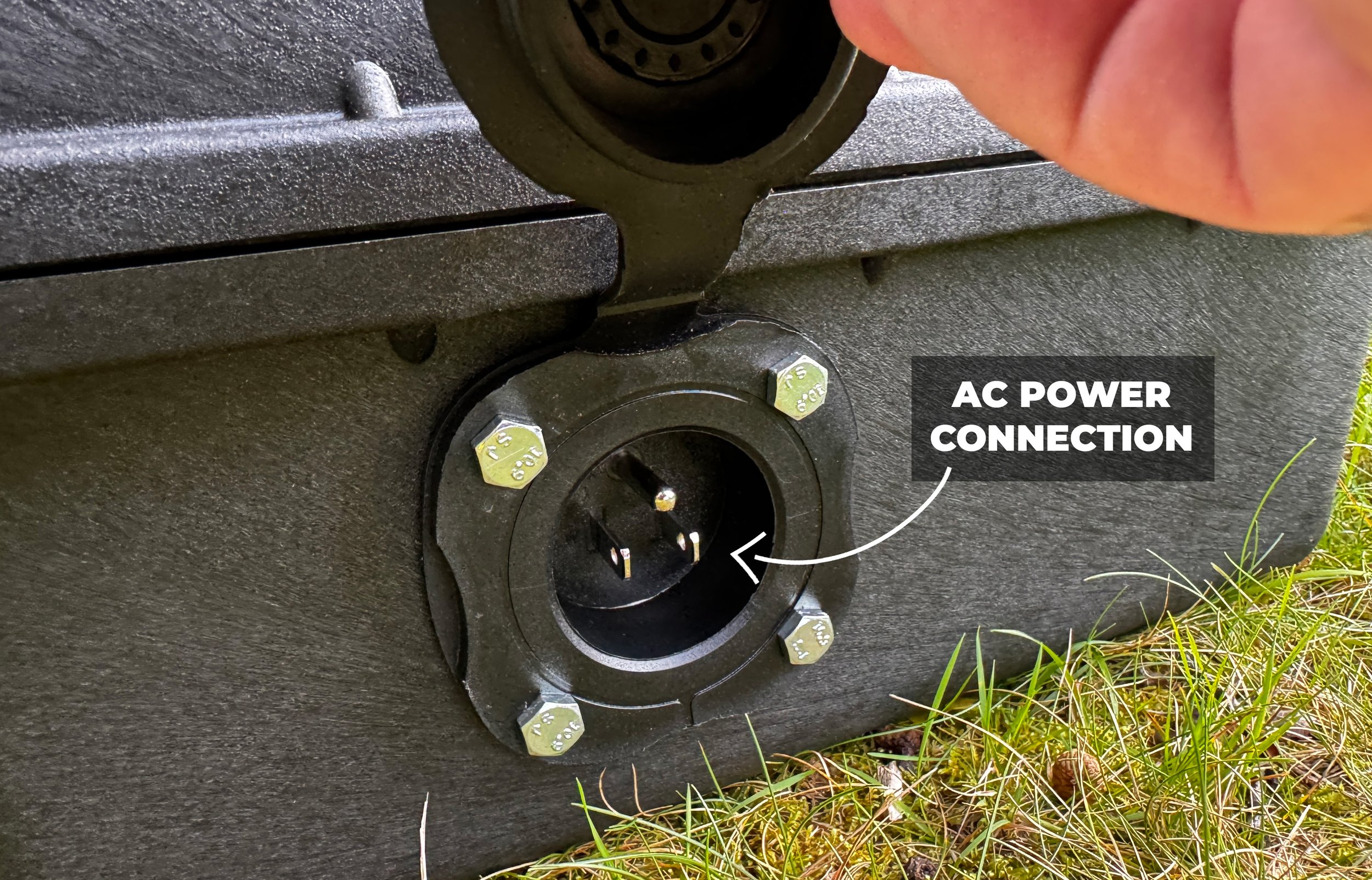 Close-up of an outdoor electrical outlet with a label pointing to it that says 'AC Power Connection'. The outlet has three prongs and is set in a black, textured surface, with some grass visible at the bottom of the photo.