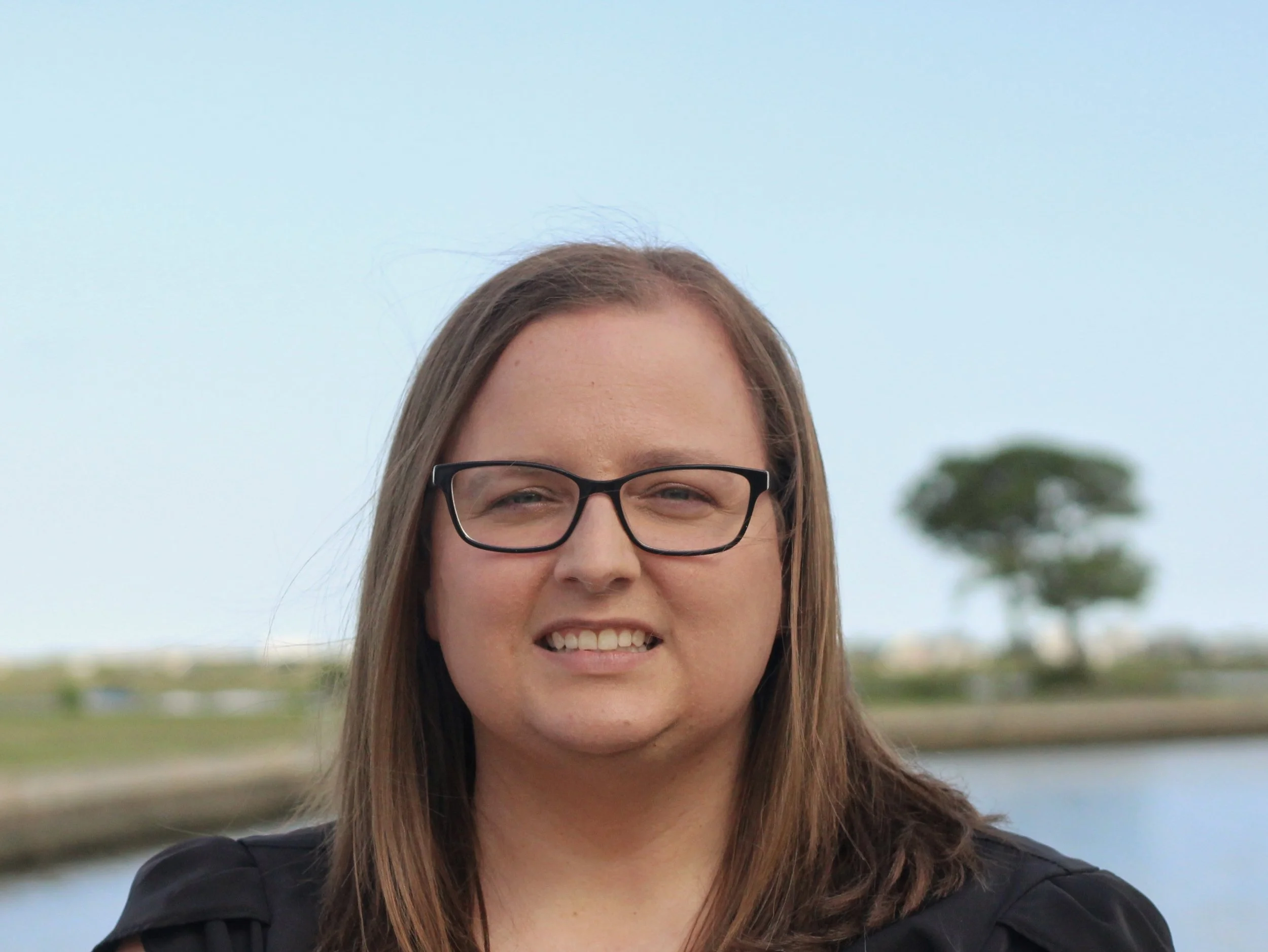 Close-up of a woman with glasses smiling outdoors, with a river and trees in the background.