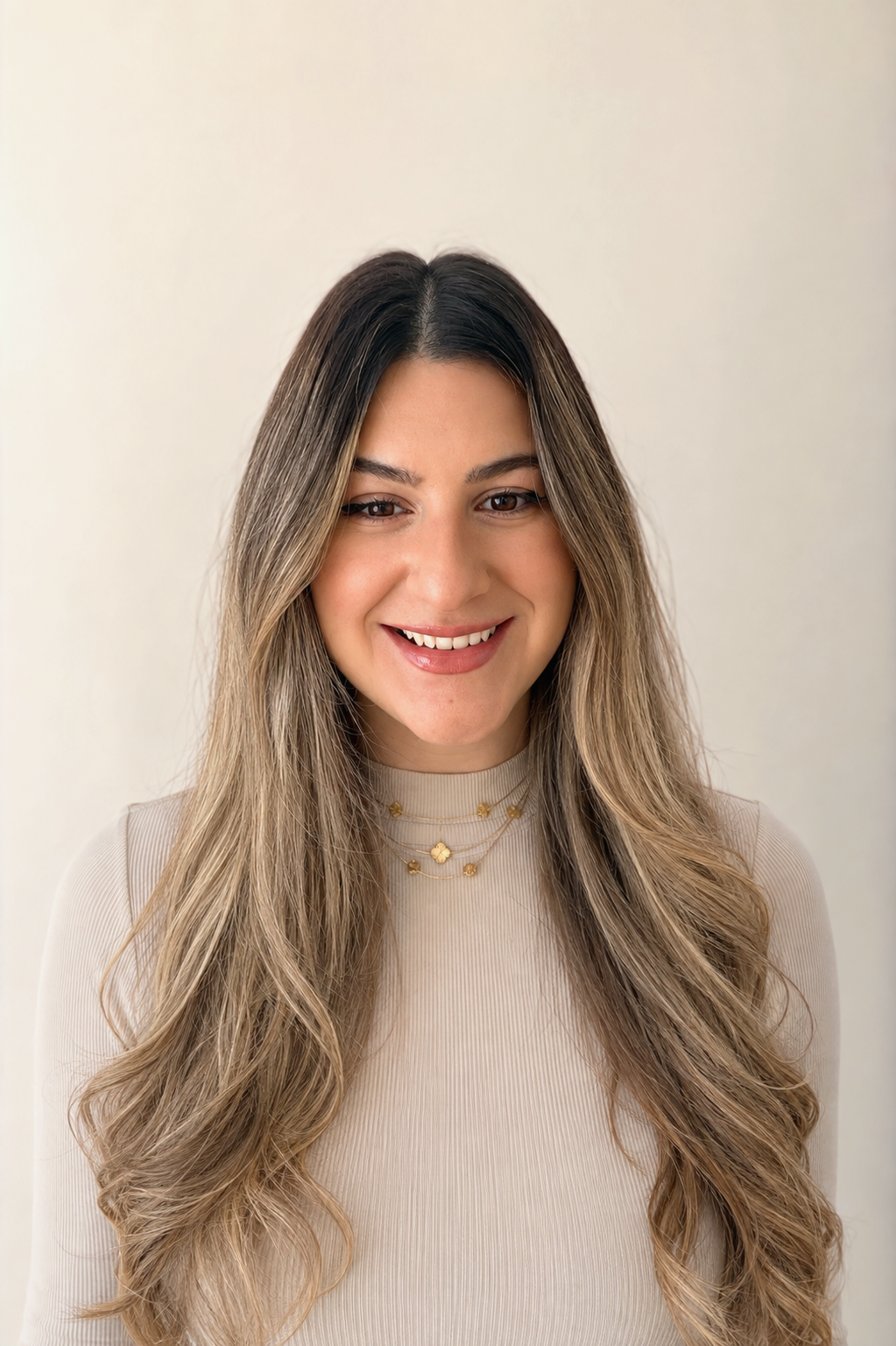 A young woman with long, wavy, ombre hair smiling, wearing a beige ribbed top and layered gold necklaces, standing against a plain off-white background.