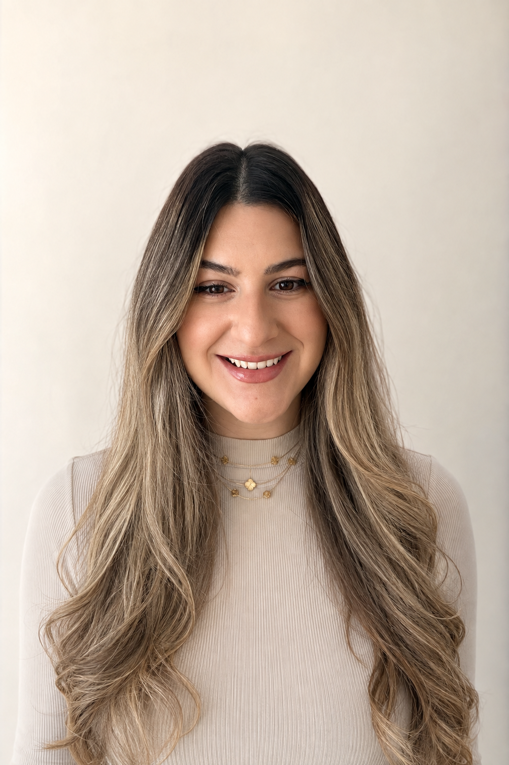 A young woman with long, wavy, blonde-brown hair, smiling, wearing a light beige top and layered gold necklaces, standing against a plain white background.