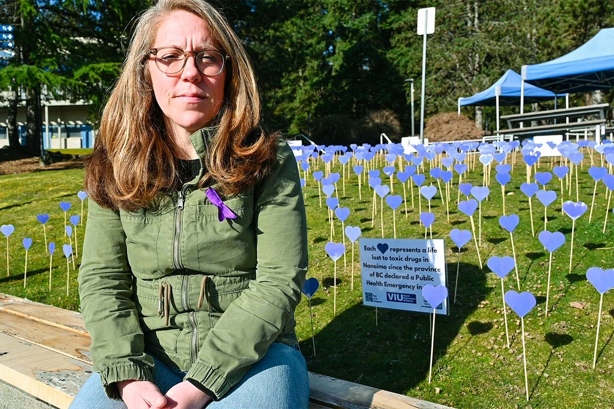 A woman with glasses and long brown hair sitting outdoors in front of a field of purple heart-shaped signs on sticks. The signs represent lives lost to toxic drugs. Two blue tents are visible in the background.