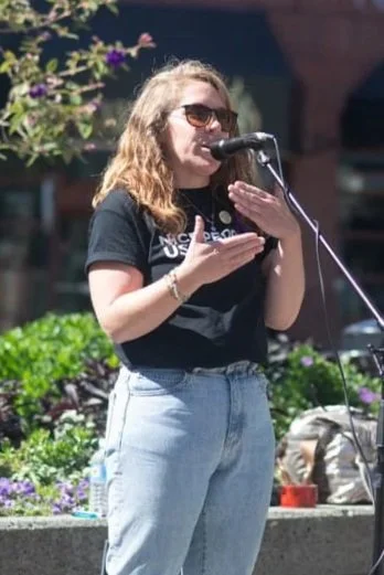 Young woman with sunglasses and wavy hair reading from a phone at an outdoor event, with plants and flowers in the background.