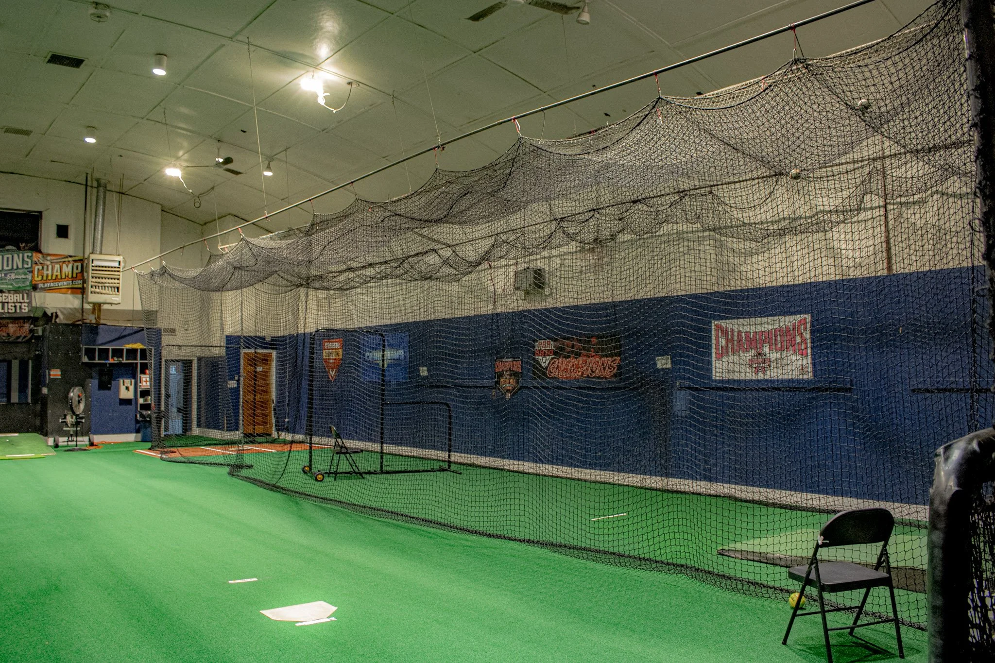 Indoor baseball training facility with green turf, a batting cage with netting, and various baseball equipment. Walls are decorated with sports banners and signs.