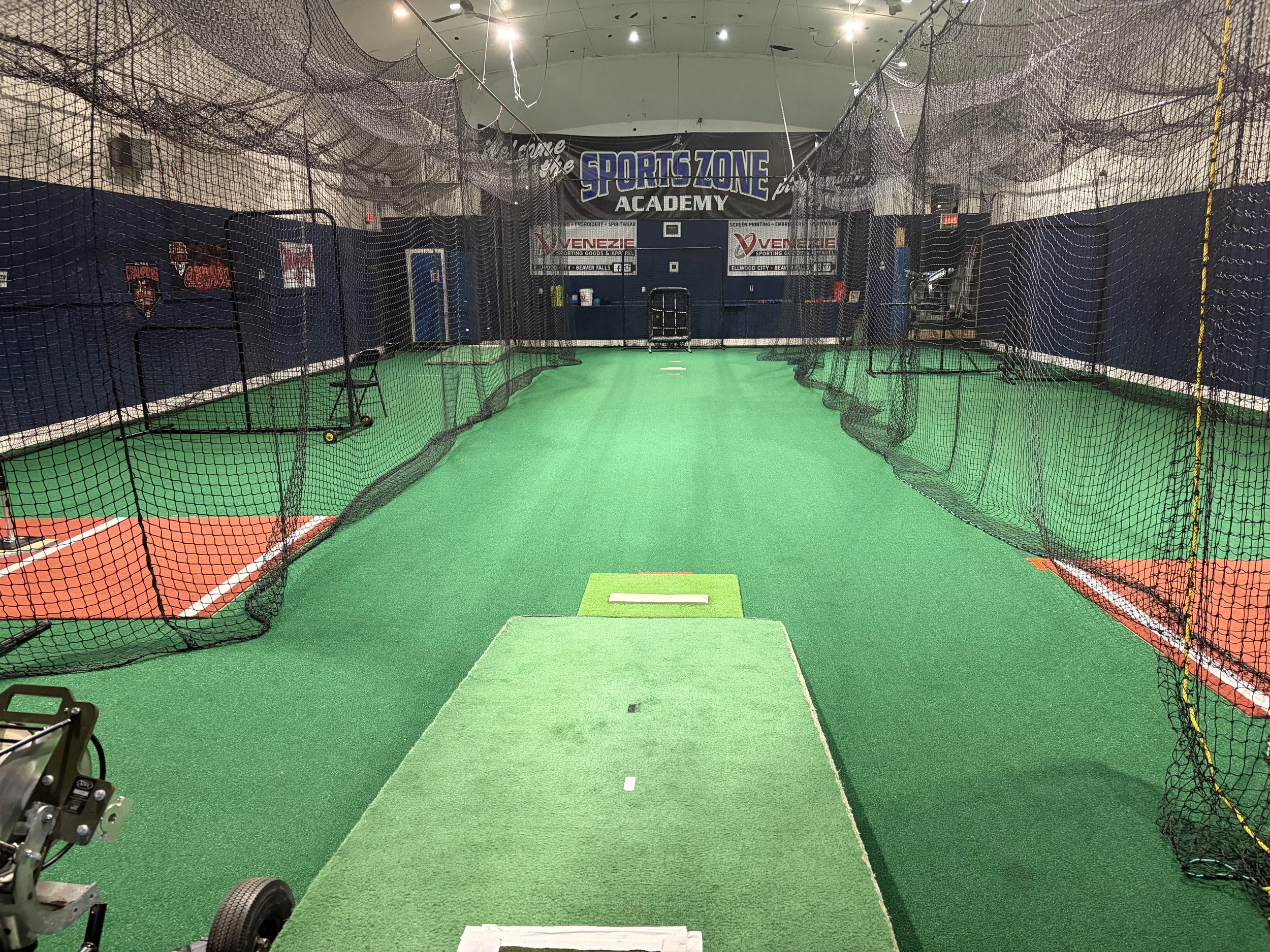 Indoor sports training area with green artificial turf, safety nets, and basketball backboard, inside a gym or sports facility.