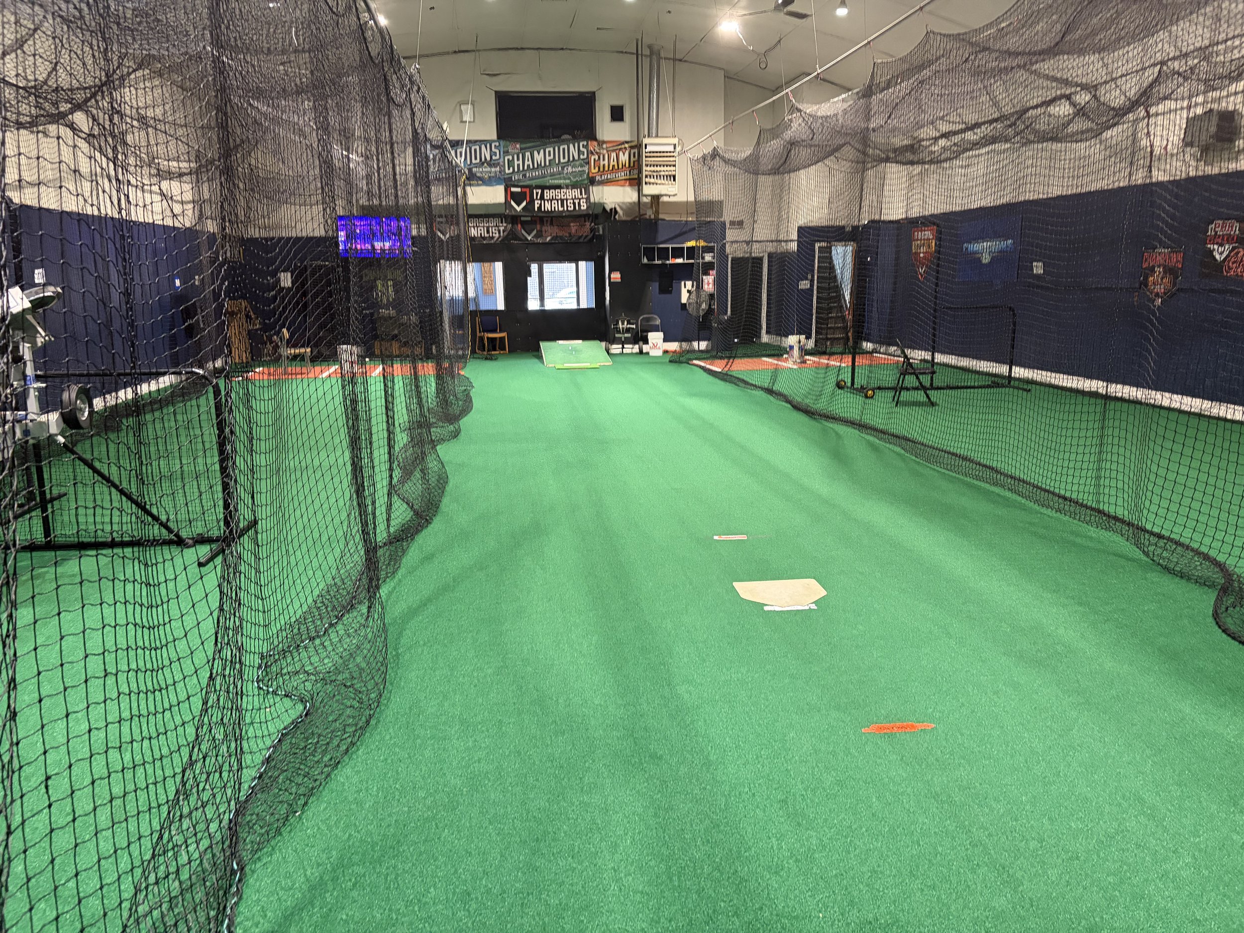 Indoor sports training facility with netted batting cages, green turf, and banners celebrating championships and baseball finalists.