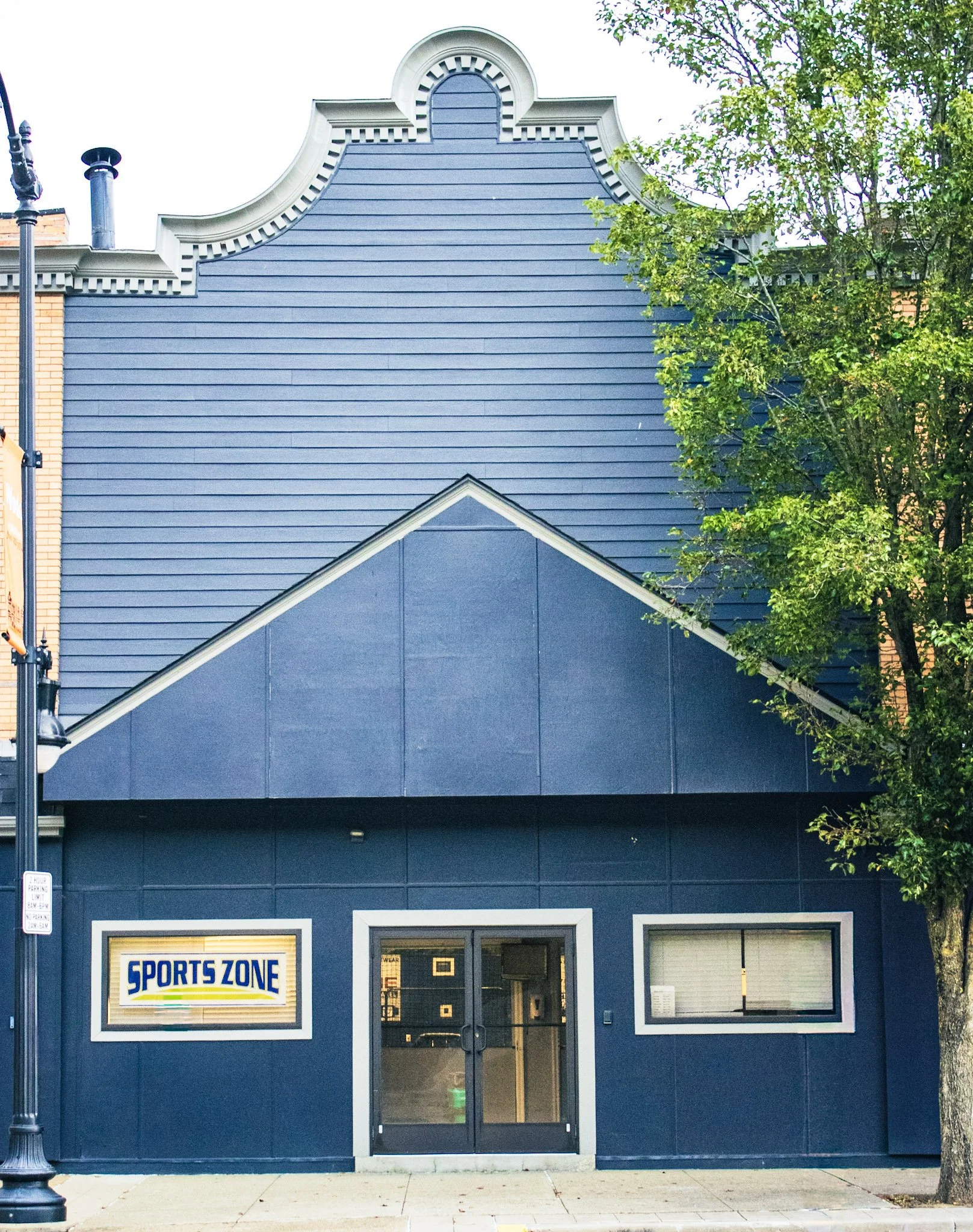 Exterior view of a blue building with a sign that reads 'Sports Zone' and windows on either side of the door. A tree partially obscures the right side of the building. A street lamp is visible on the left.