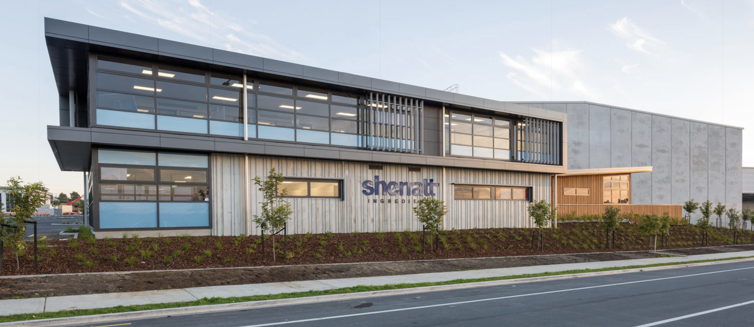 Modern industrial building with large glass windows, a sign reading 'Sheratt Ingredients', surrounded by planted greenery and a paved street in the foreground.