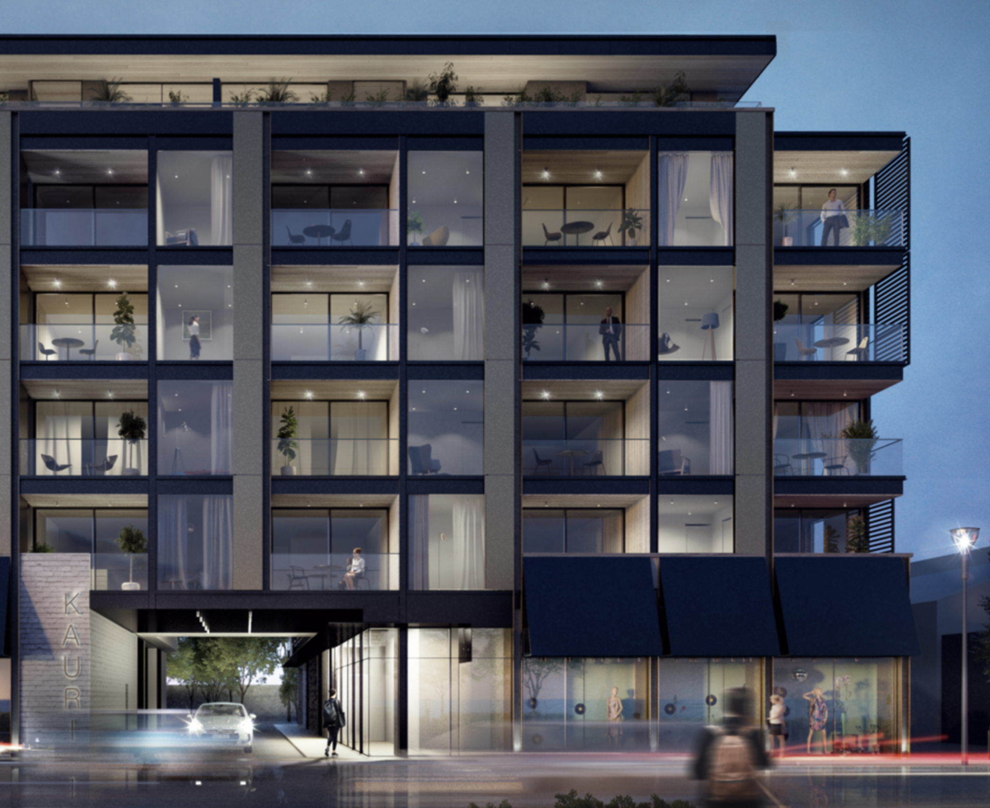 Nighttime exterior view of a modern multi-story building with large glass windows, some with interior lighting and furniture visible. Several people can be seen through the windows, and a woman is standing on a balcony to the right. Trees and a pathway are visible at the base of the building.