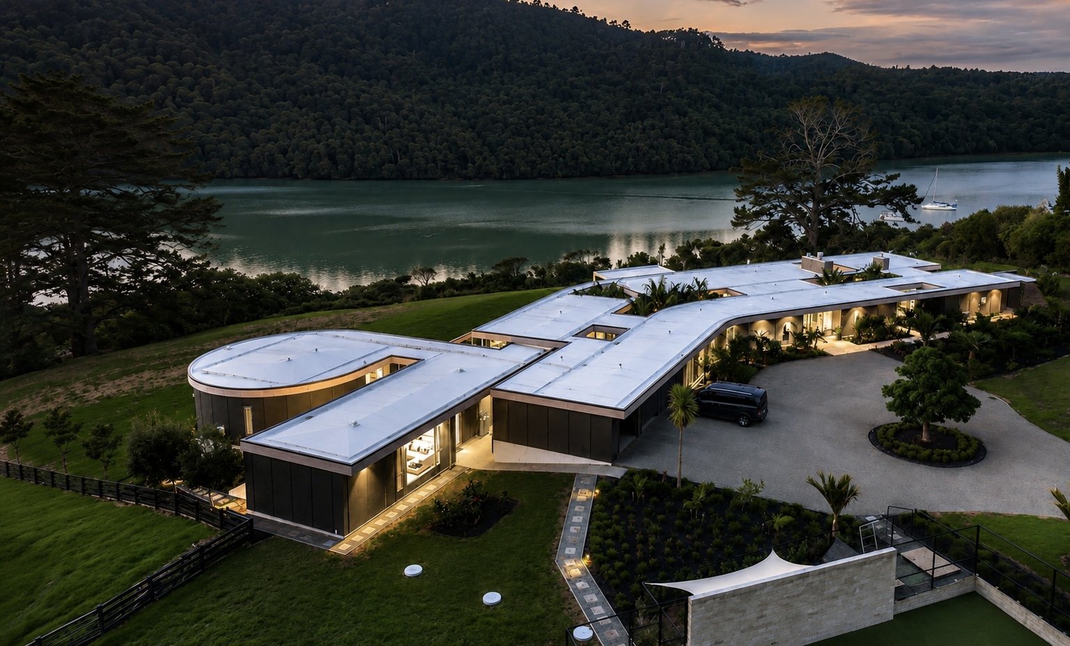 A modern house with a white roof and large glass windows, surrounded by green lawns, trees, and a view of a lake and hills at sunset.