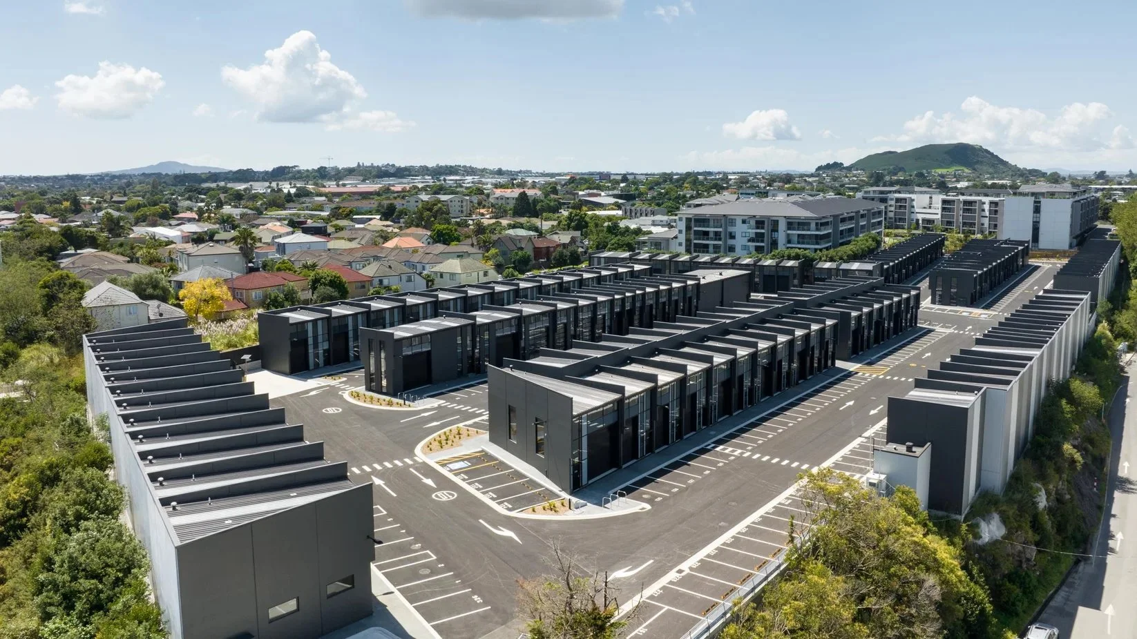 A modern, multi-level commercial or residential building complex with a large parking lot, surrounded by a suburban neighborhood with trees and houses, and a hilly landscape in the background under a partly cloudy sky.