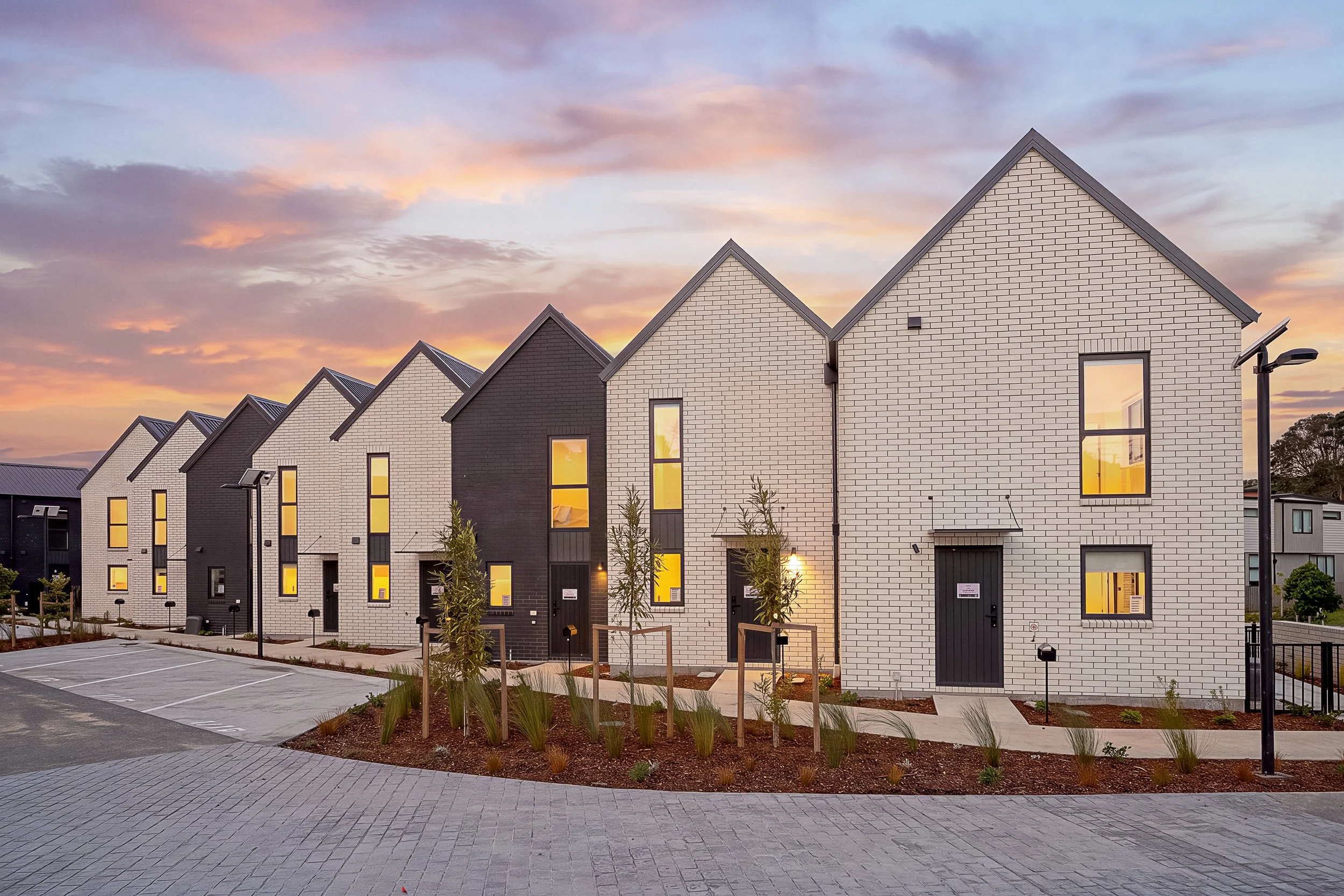 Modern townhouse row with white and black facades under a colorful sunset sky, with small trees, streetlights, and parking spots in the foreground.