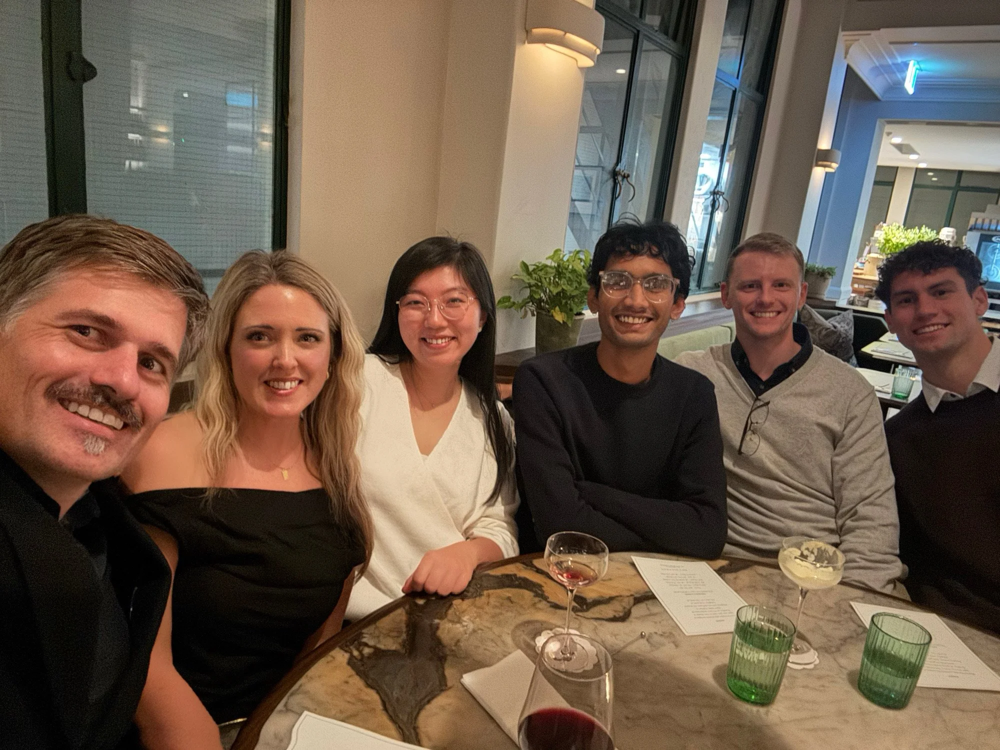 Group of five people smiling and sitting together at a restaurant table with drinks and a menu on the table.