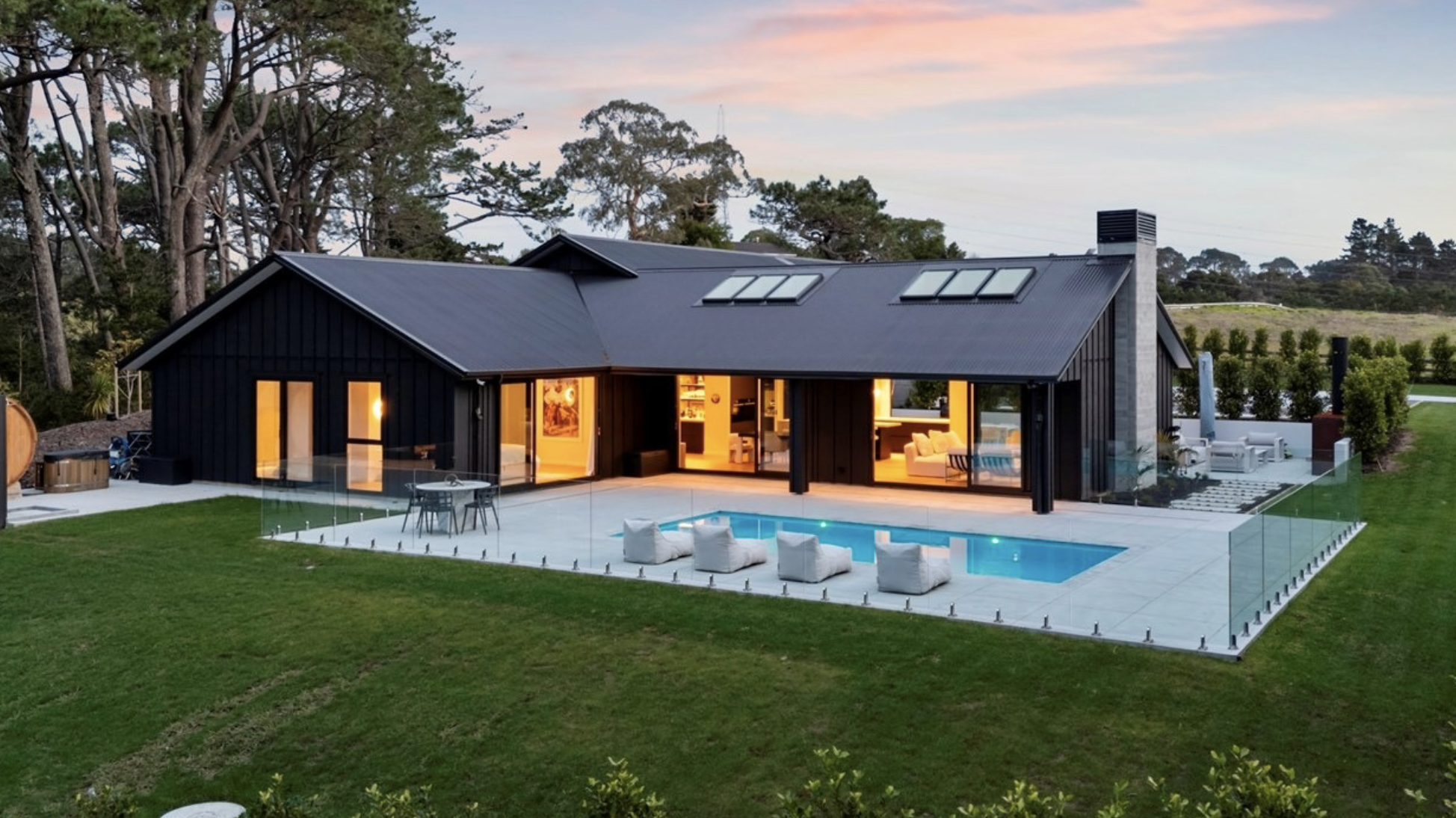 Modern black house with a sloped roof, large glass doors, and skylights, surrounded by a lush yard with trees in the background. The house features an outdoor patio area with seating, a small swimming pool, and a glass fence at the backyard.
