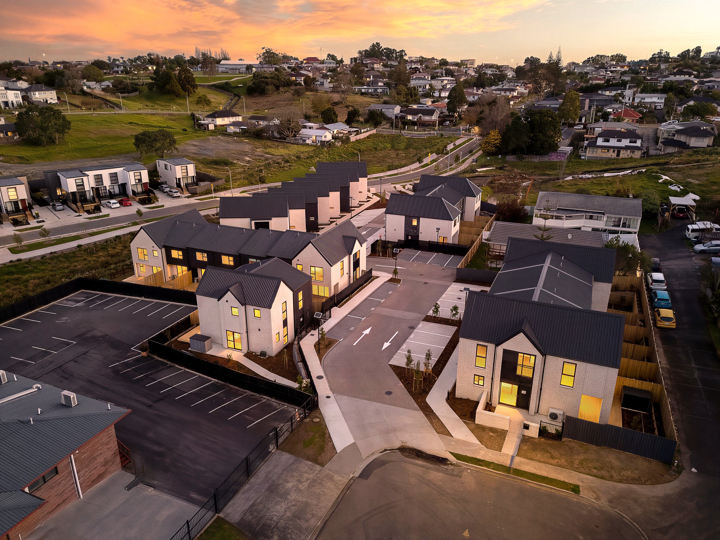 Aerial view of a suburban neighborhood at sunset, showing modern houses with dark roofs, parking lots, and green yards.