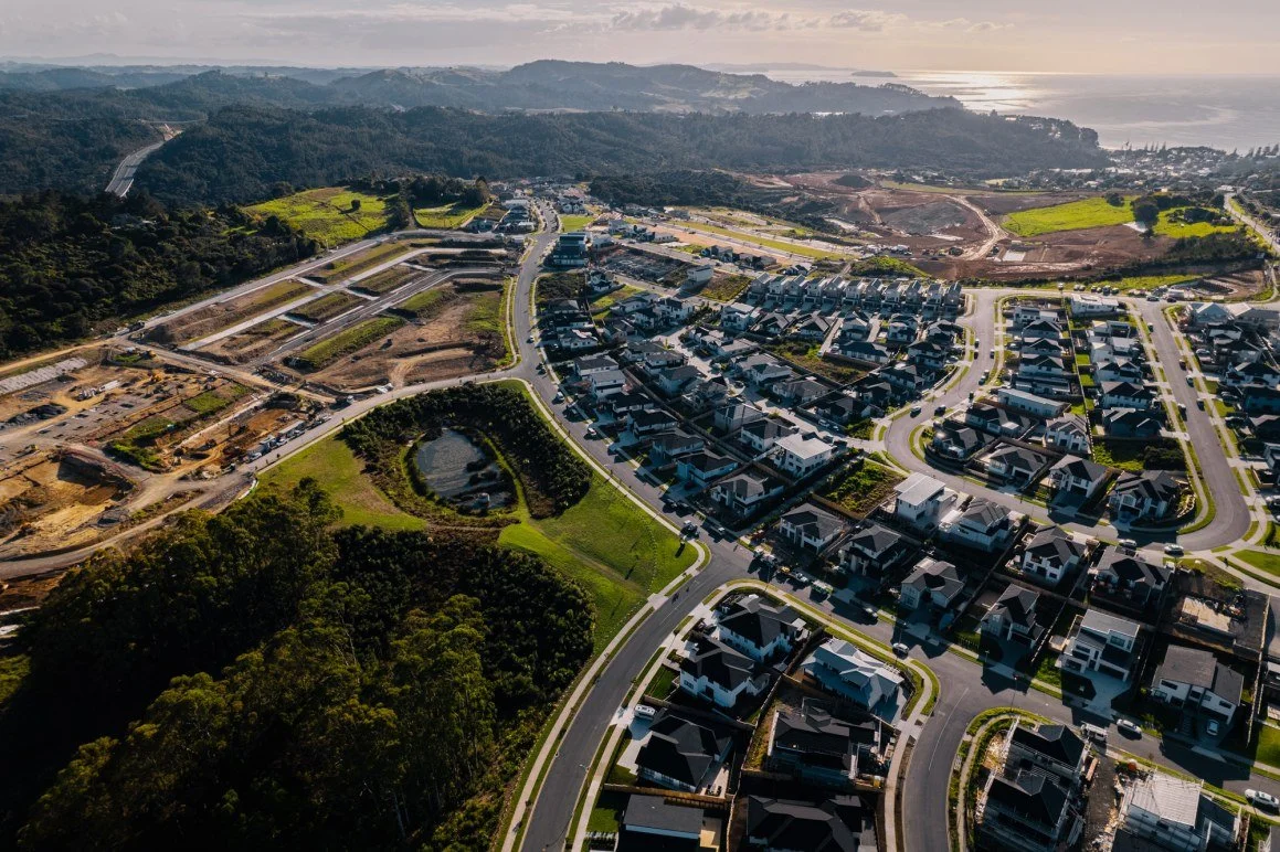 Aerial view of a residential neighborhood with new houses, winding streets, green lawns, and nearby undeveloped land and hills, with a distant body of water in the background.