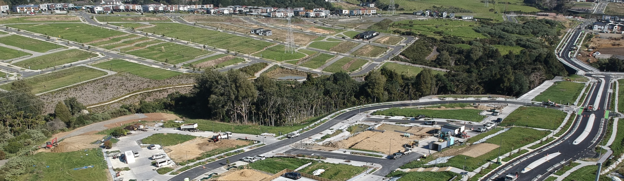 An aerial view of a residential development area under construction, with newly paved roads, construction vehicles, and some completed homes surrounded by green spaces and trees.